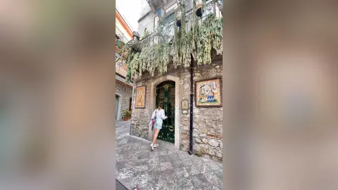 Stylish woman in a white shirt and denim shorts walking through a charming, sunlit European cobblestone alley.