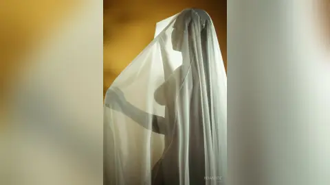 KeianHoz stands in profile behind a sheer white veil that covers her head and body, revealing the silhouette of her naked chest and torso.