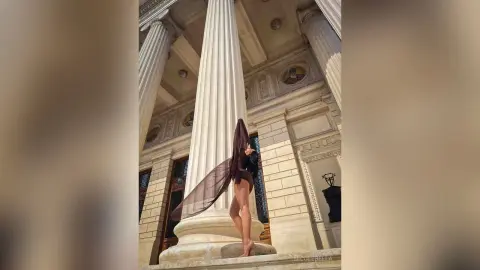 NicoleBella stands against a large white column wearing a sheer black mesh bodysuit that shows her bare skin.