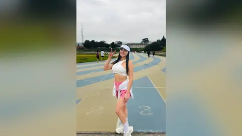 Cheerful young woman in white crop top and pink shorts posing on a blue running track under a cloudy sky.
