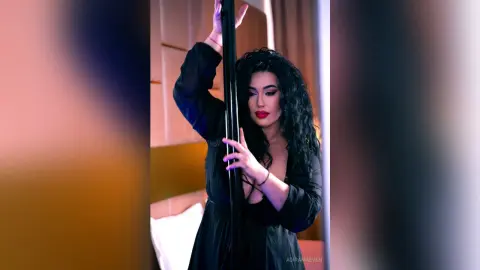AdiraMaeven poses with a black pole, wearing a black long-sleeve top with her long, dark curly hair down.