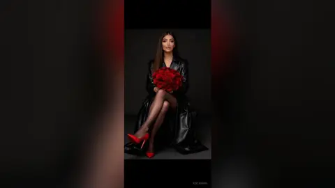 Elegant woman in a black leather coat and red heels holding a large bouquet of red roses against a dark background.