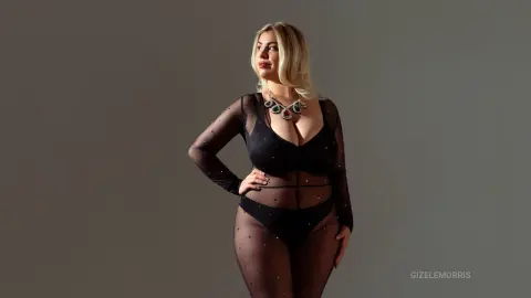Blonde woman in a black sheer bodysuit and statement necklace posing against a grey studio background.