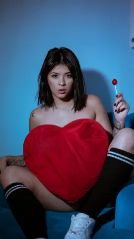 Young woman in black thigh-high socks holding a red heart pillow against a blue background with moody lighting.
