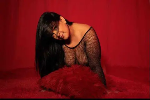 Smiling woman in black fishnet top posing against a red backdrop for a glamorous portrait.