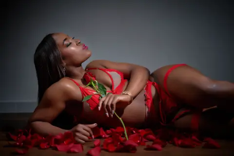 Elegant woman in red lace lingerie lying on a floor covered in rose petals in a moody, romantic setting.