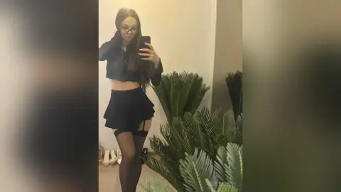 Young woman in a black sheer blouse and mini skirt taking a mirror selfie in a modern room.