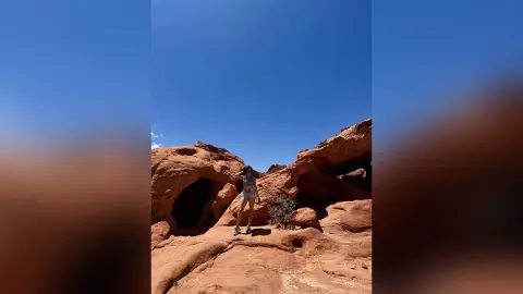 Young woman in casual summer outfit standing by red desert rock formations under a clear blue sky.