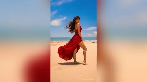 Elegant woman in a flowing red gown posing on a sunny beach under a clear blue sky.
