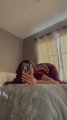 Woman in a red patterned loungewear set taking a mirror selfie on a floral bed in a sunlit bedroom.