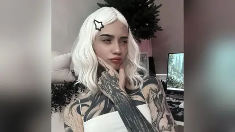 Tattooed woman with long white hair in a white tank top posing thoughtfully in a cozy indoor setting.