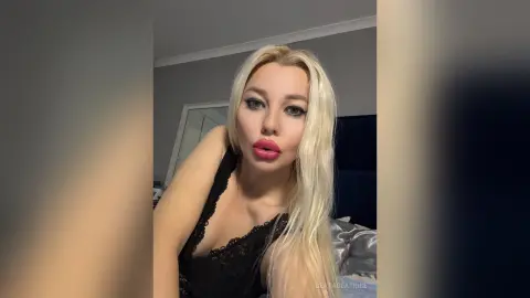Blonde woman in a black lace top posing with a glamorous look in a modern bedroom setting.