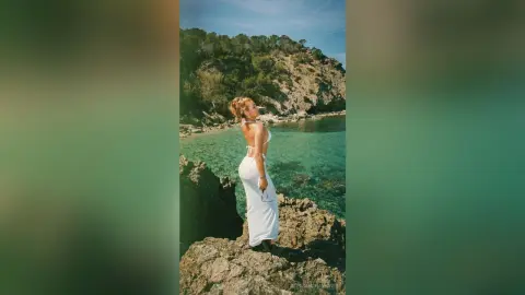Blonde woman in a white summer dress standing on a rocky cliff overlooking a turquoise Mediterranean sea.