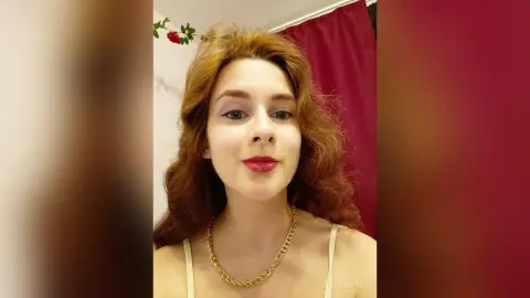 Smiling woman with wavy red hair wearing a gold chain necklace in a warm indoor setting.