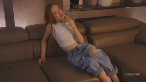 AddieRunk poses while reclining on a brown leather couch, wearing a white crop top and ripped denim shorts with her long red hair down.