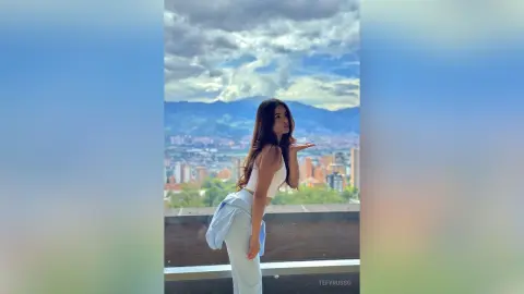 TefyRusso poses outdoors against a mountain and city view, wearing a white tank top and light blue pants with a light blue shirt tied around her waist.