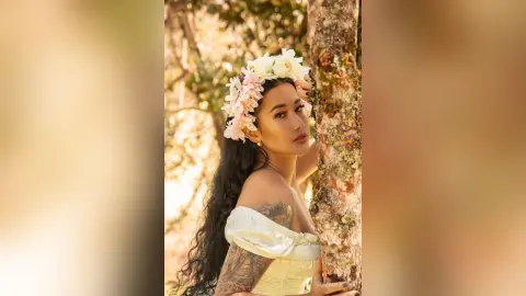 JuliahLewis poses outdoors against a tree trunk wearing a white off-the-shoulder top and a white flower crown in her long dark hair.