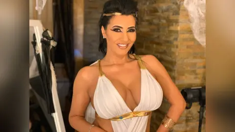 MissyJolie smiles wearing a white plunging halter-neck top with gold trim and a gold headband.
