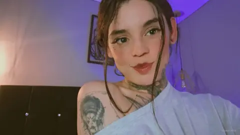 Young woman with tattoos in a white top posing in a room with moody purple lighting.