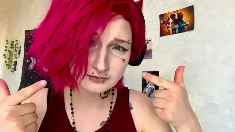 Woman with bright pink hair and facial piercings wearing a maroon tank top in a cozy indoor setting.