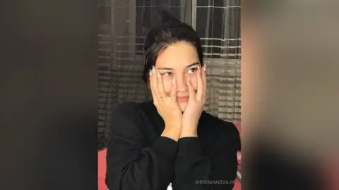 Young woman in a black sweater resting her face on her hands in a cozy indoor setting with a thoughtful mood.
