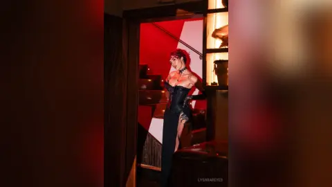 LysaraReyes poses in a black corset and long skirt, standing in a red-lit room next to a dark wooden staircase.