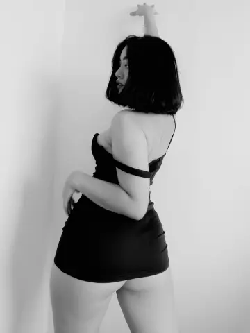 AnnaKomatsuu poses in a black bodysuit, looking over her shoulder with her arm raised against a plain white wall.