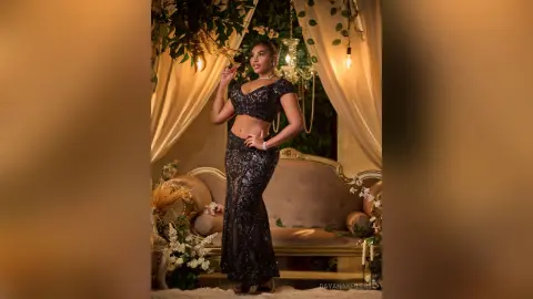 DayanaKeilers poses standing in a black sequined two-piece outfit against a decorative sofa and white drapes.