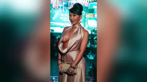 Dolce poses in a tan satin halter dress with a deep plunging neckline, showing cleavage, while holding a gold clutch.