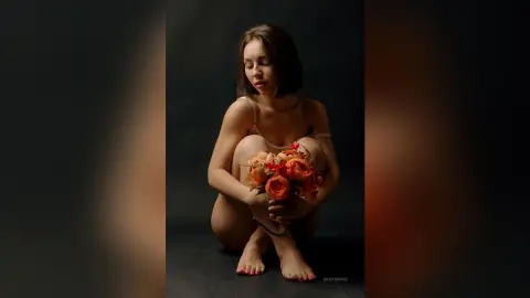 Serene woman in minimal attire holding a vibrant orange flower bouquet against a dark moody background.