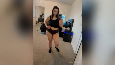 Curvy woman in black tank top and maroon shorts taking a mirror selfie in a cozy bedroom setting.