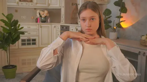 Young woman in a white shirt and tank top sitting in a bright, modern kitchen with indoor plants.