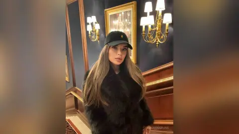 Stylish woman in a black faux fur coat and baseball cap posing in a luxurious, dimly lit room.