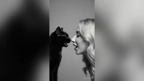 Black and white portrait of a blonde woman smiling at a black cat in a minimalist studio setting.