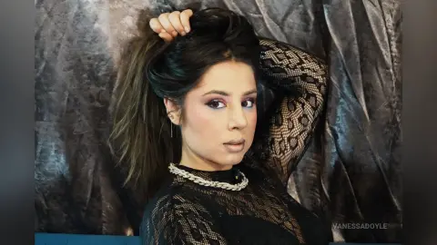 Glamorous woman in a black lace long-sleeve top posing against a textured grey backdrop with elegant makeup.