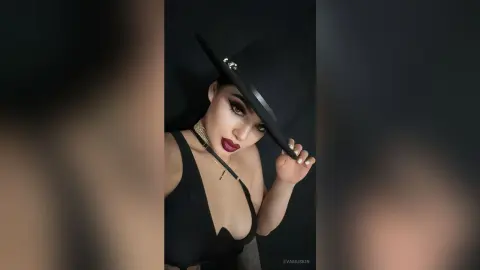 Glamorous woman in a black tank top and wide-brimmed hat posing with elegant makeup against a dark background.