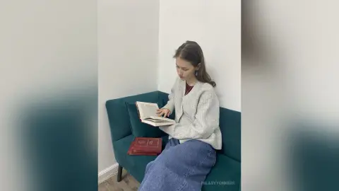Young woman in a cozy grey cardigan reading a book on a teal velvet sofa in a bright minimalist room.
