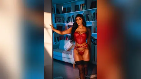Glamorous woman in red lace lingerie posing in a sunlit modern living room with bookshelves.
