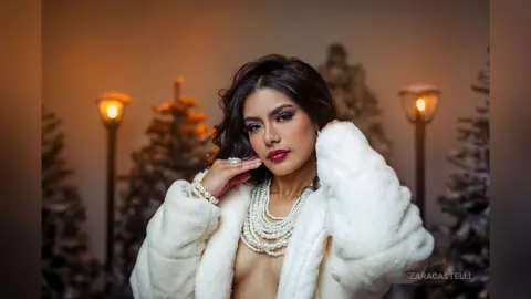 Glamorous woman in a white faux fur coat and pearl necklace posing in a festive holiday setting.