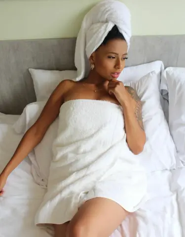 Relaxed young woman wrapped in a white towel and head wrap sitting on a bed in a cozy bedroom setting.
