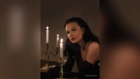Elegant woman in a black dress posing by glowing candles in a moody, romantic dimly lit setting.