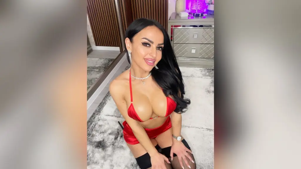 EmilyMae wears black glasses and a red sheer mesh outfit with a shiny red bra and skirt, posing on a black couch with long black hair.