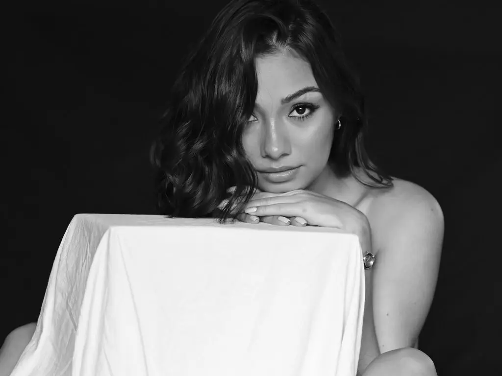 Moody black and white portrait of a young woman leaning on a white draped surface against a dark background.