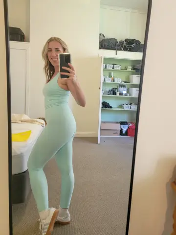 Smiling woman in mint green yoga set taking a mirror selfie in a bright bedroom.