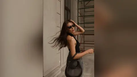 Stylish woman in a black sleeveless top and sunglasses posing outdoors against a modern building wall.