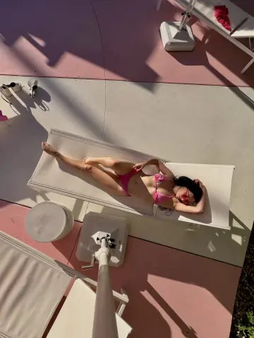 Young woman in a pink bikini relaxing on a white lounge chair at a sunny outdoor poolside patio.