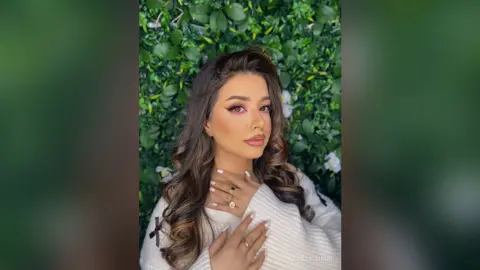 Woman with glamorous makeup and wavy hair posing against a lush green leaf wall in a serene setting.