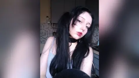 Young woman with long black hair and dark makeup wearing a grey tank top in a moody indoor setting.