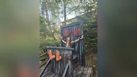 Woman in a dark outfit posing playfully behind a spooky Dracula themed wooden prop in a lush forest setting.