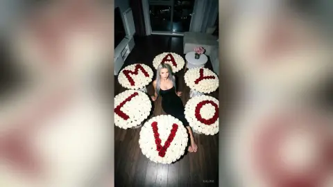 Kalira poses on a dark floor surrounded by large white flower arrangements shaped like the letters in 'I LOVE MAY', wearing a black dress.